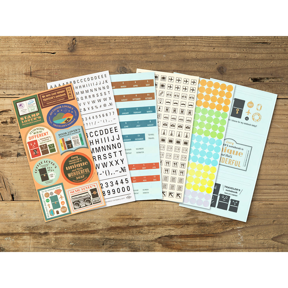 A set of colorful planner sticker sheets arranged on a rustic wooden tabletop. The stickers feature retro-style labels, alphabet letters, icons, and decorative text in teal, orange, yellow, green, and black.