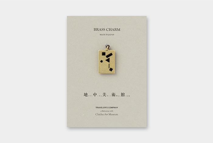 A brass charm pendant with black geometric accents, presented on a light beige card labeled 'BRASS CHARM' with additional text below, photographed against a light gray background.