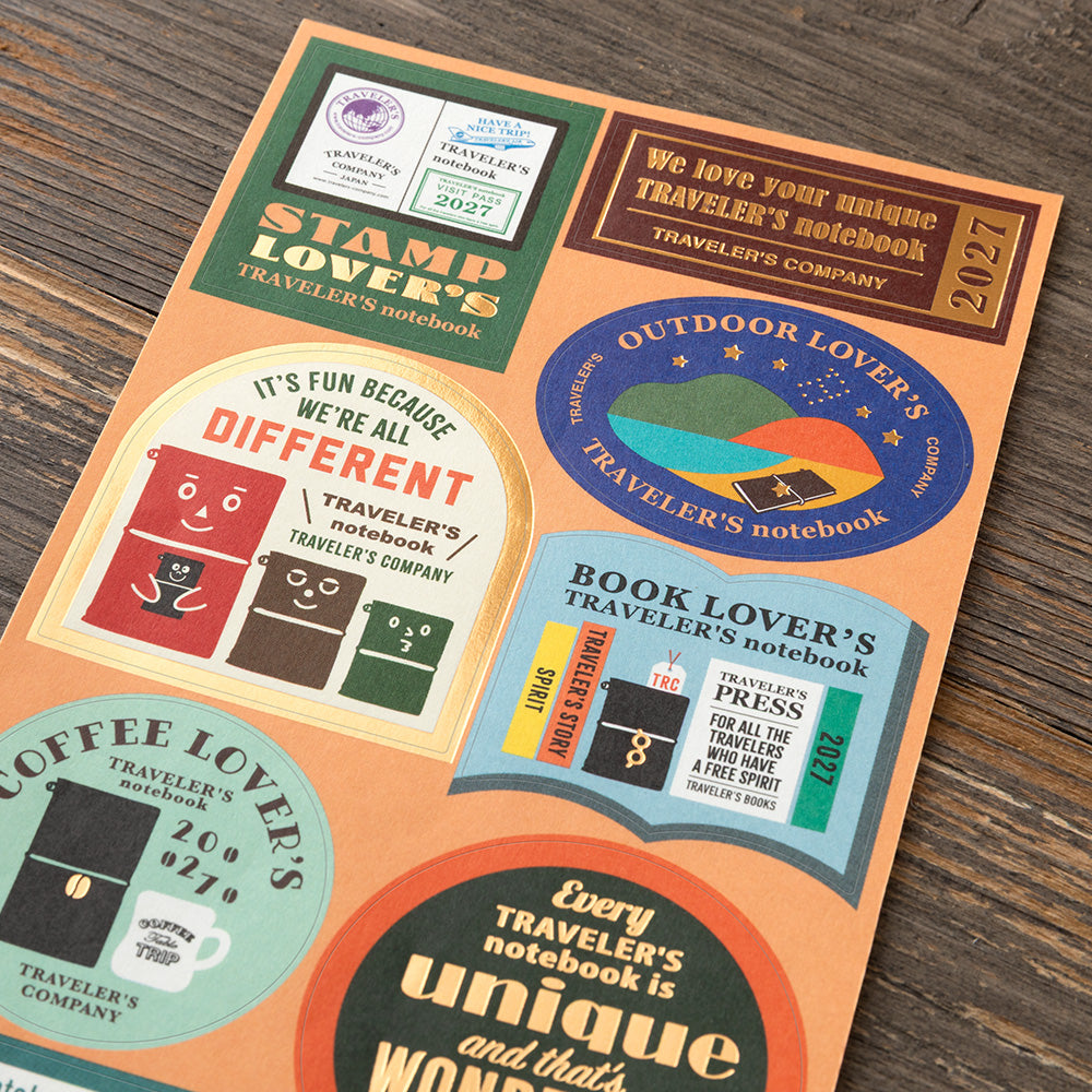 A flat-lay of a colorful printed sticker sheet featuring various travel-themed circular and rounded decals with slogans like 'Stamp Lover's' and 'Outdoor Lover's' on a dark wood background.