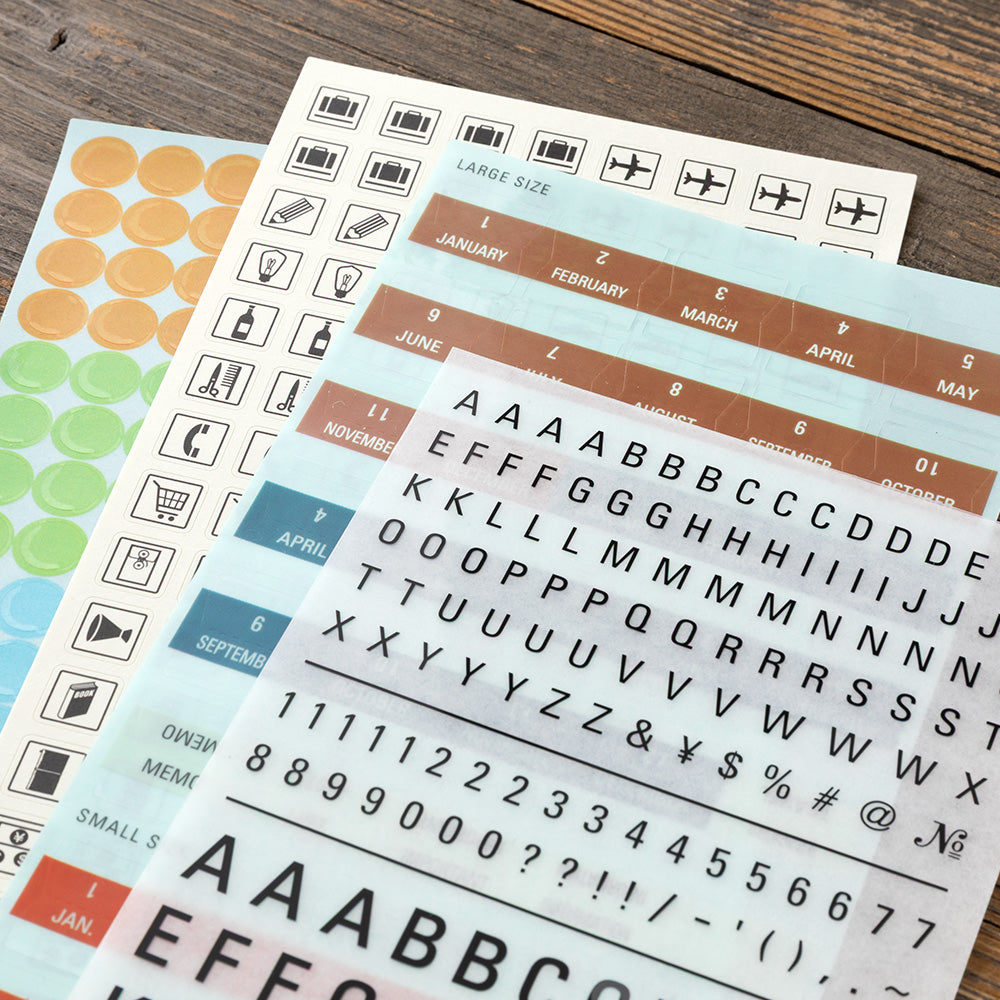 Planner sticker sheets laid out on a wooden surface, featuring travel icons, colored date blocks, and alphabet stickers.