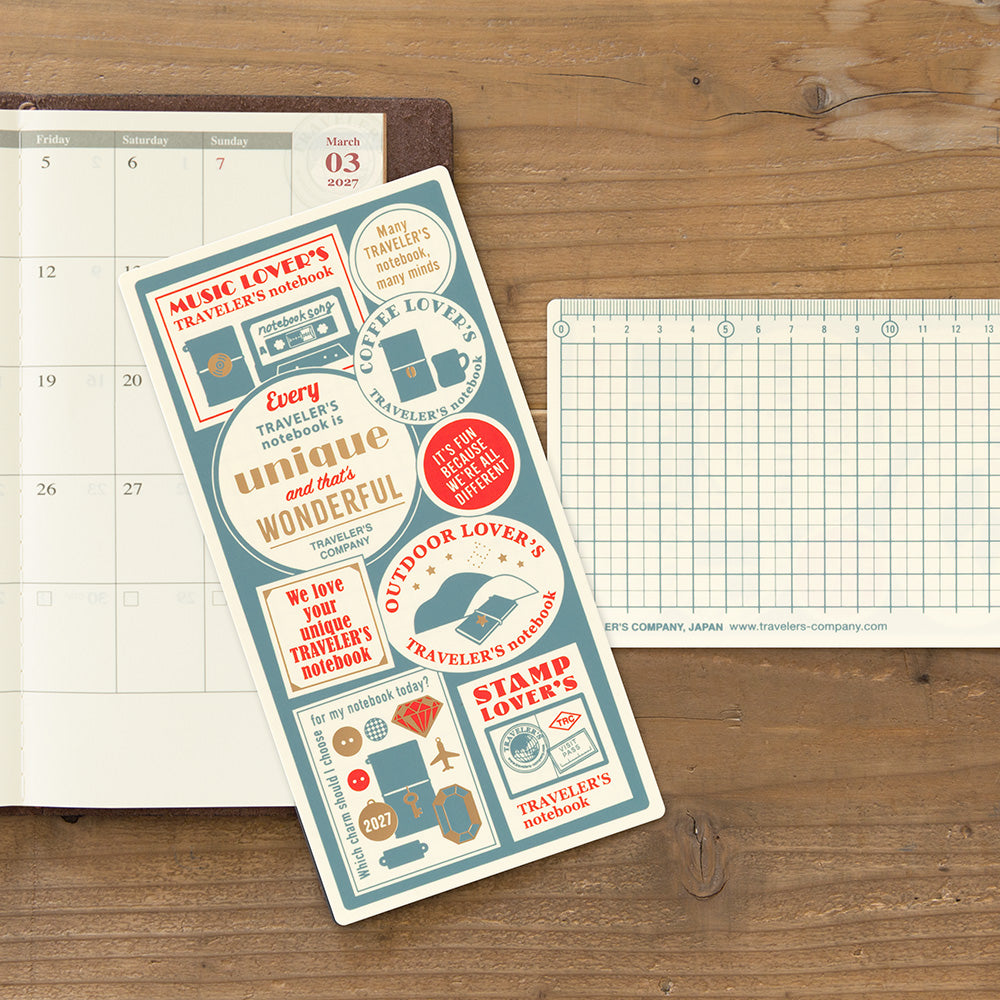 A sheet of decorative planner stickers with vintage travel-themed designs and text, displayed on a wooden tabletop next to a calendar page and a graph paper sheet.