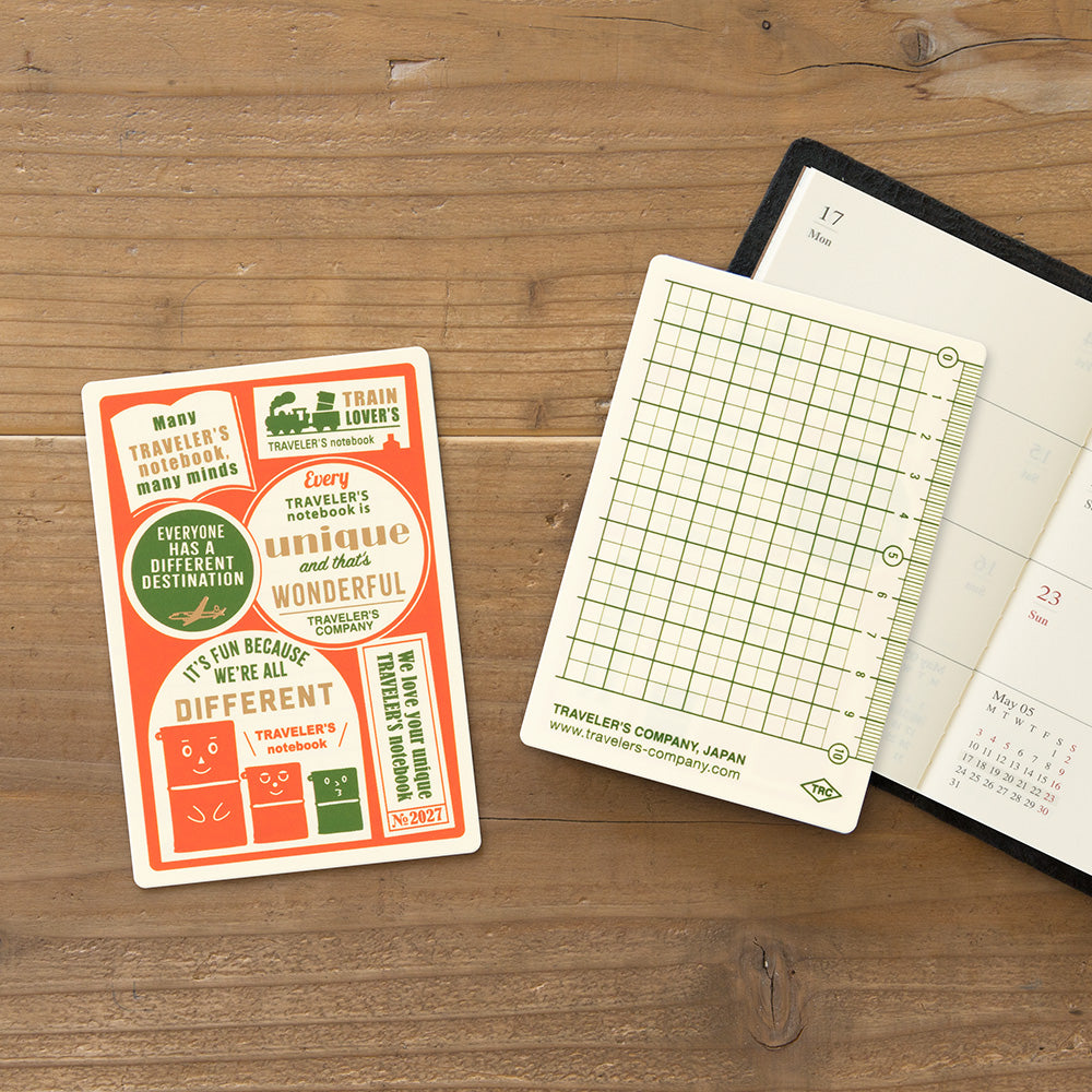 A set of Traveler's Company stationery items arranged on a wooden tabletop, including a printed card with retro travel-themed graphics and a white grid notepad partially tucked under an open black planner.
