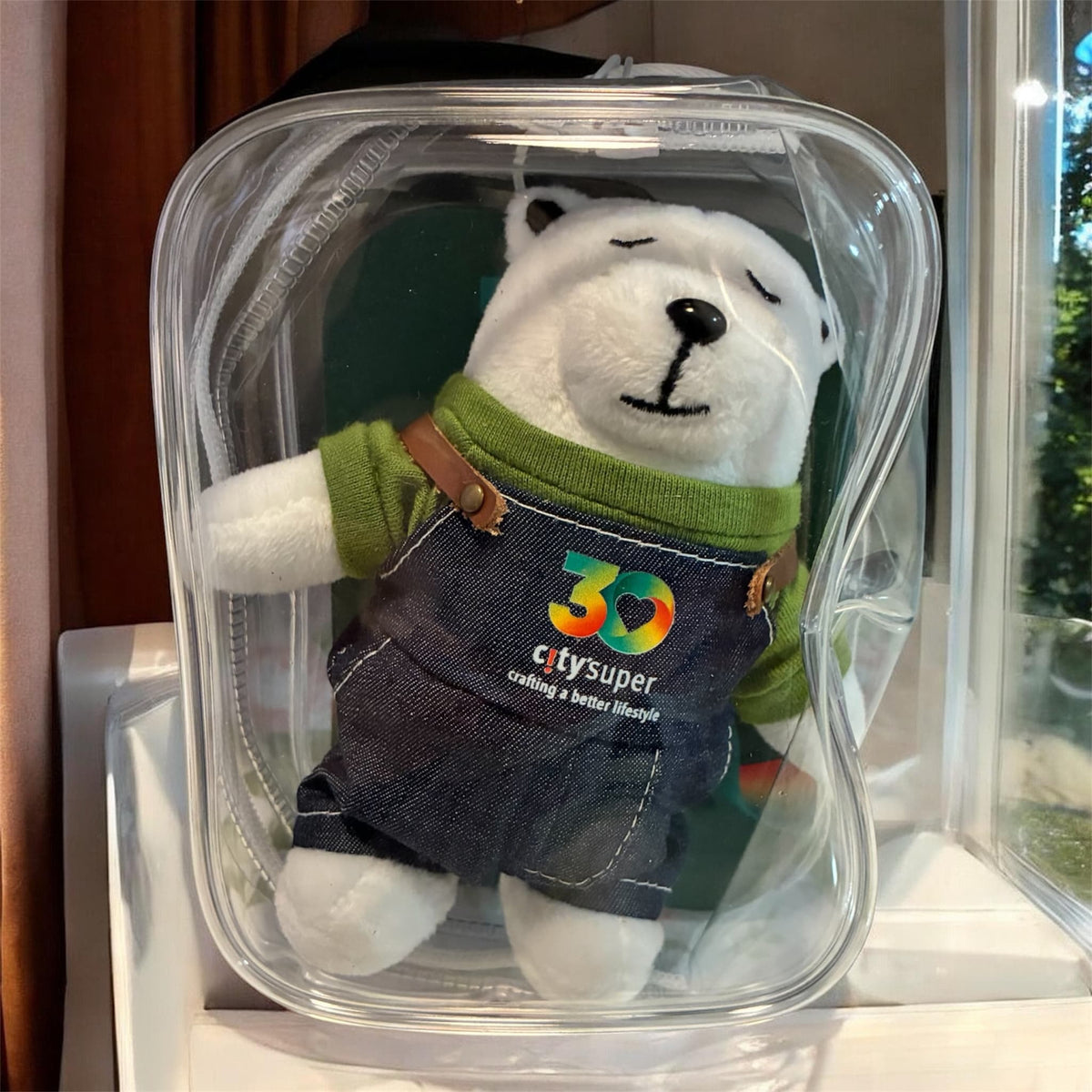A white polar bear plush toy wearing a green shirt and dark denim overalls with a colorful '3D citysuper' logo, packaged inside a clear plastic bag.