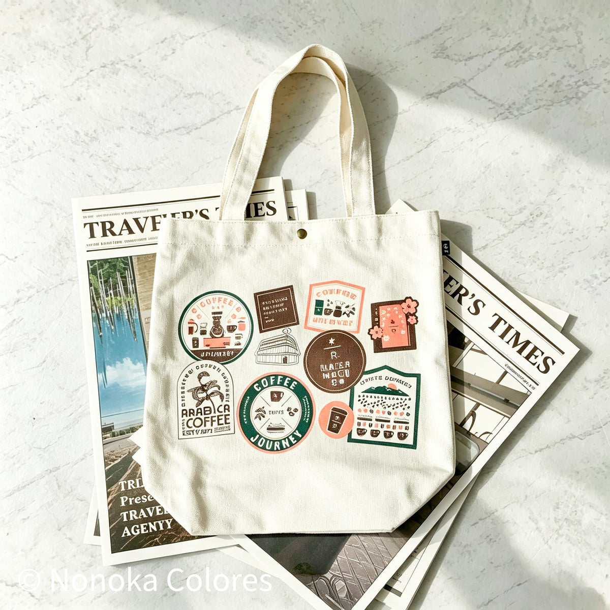 A cream canvas tote bag with a colorful coffee-themed sticker print, styled on top of vintage travel magazines on a light marble surface.