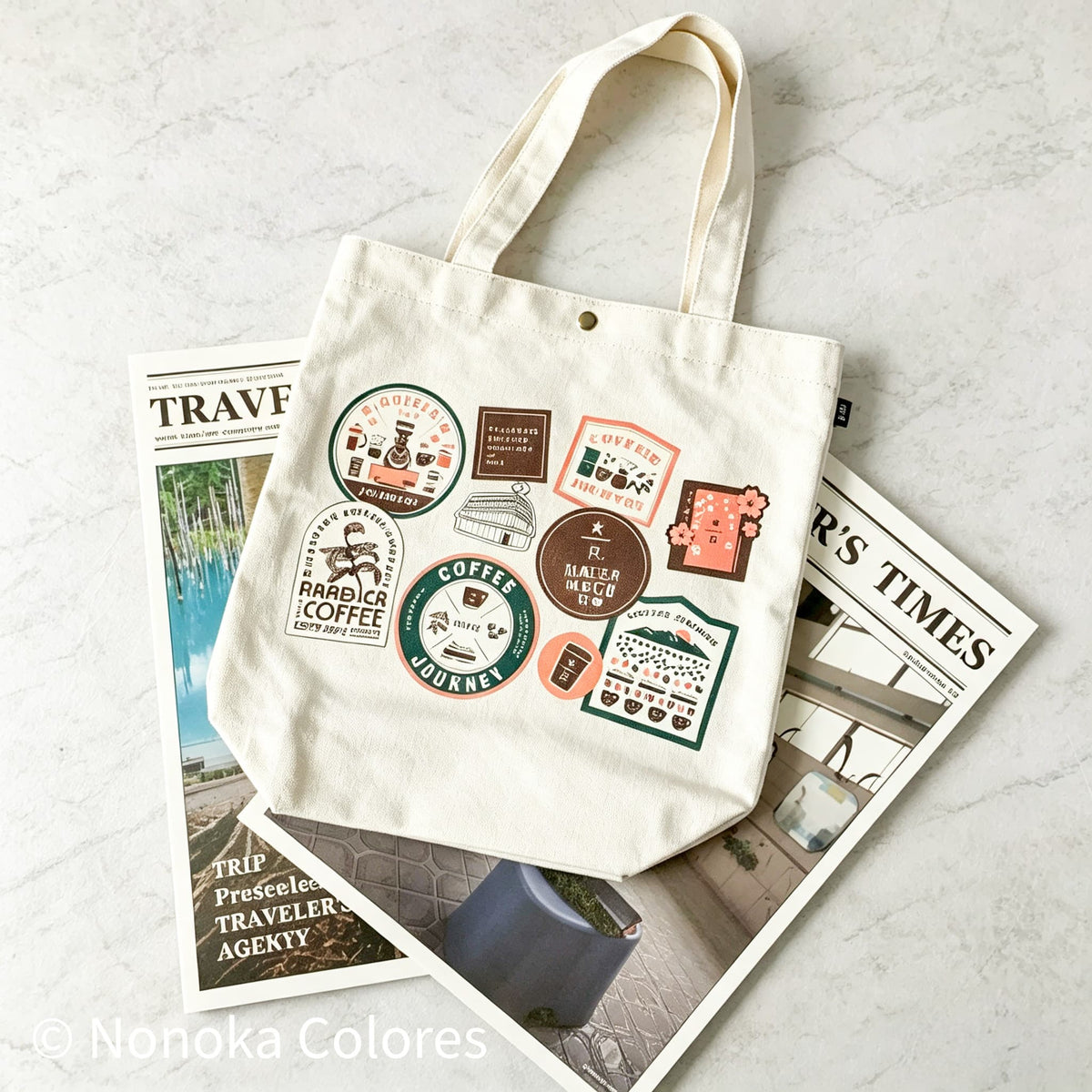 An off-white canvas tote bag with a multicolor collection of coffee-themed travel stickers, styled on top of two vintage-style travel magazines on a light gray marble surface.