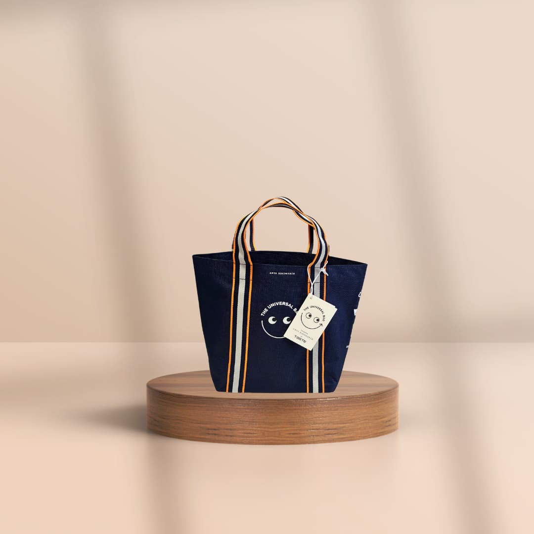 A navy blue canvas tote bag with striped handles and a white hanging tag, displayed on a round wooden pedestal against a warm beige background.