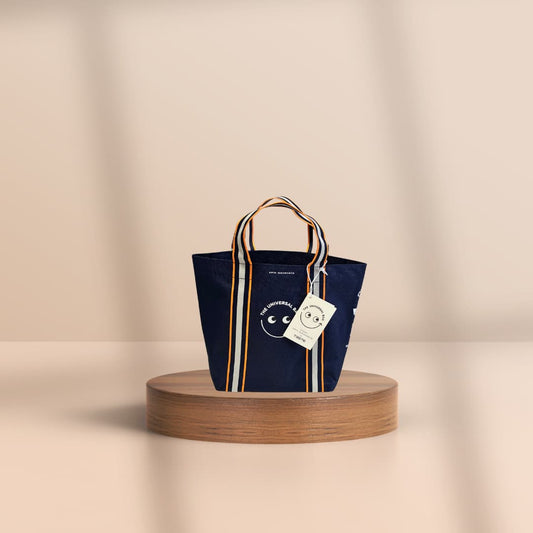 A navy blue canvas tote bag with striped handles and a white hanging tag, displayed on a round wooden pedestal against a warm beige background.