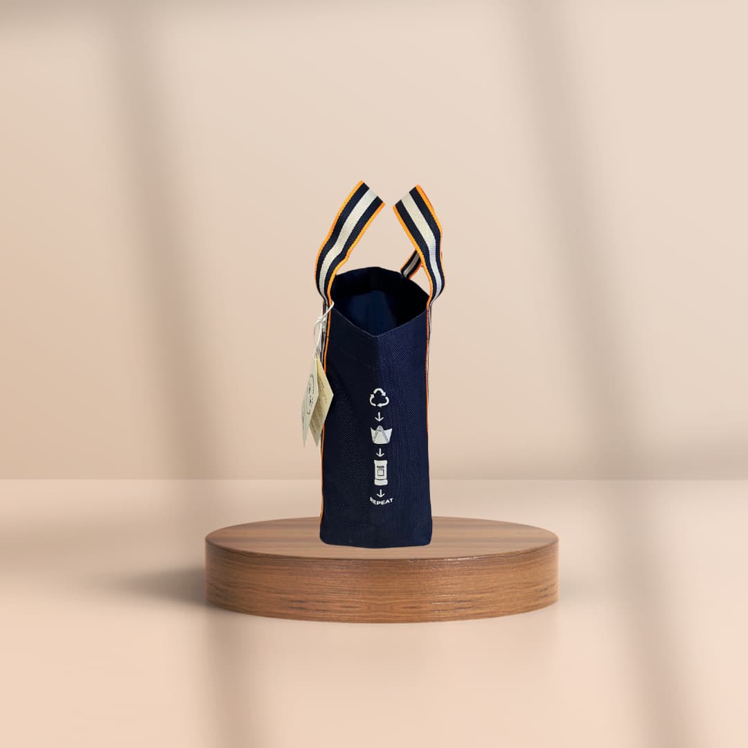 A navy blue fabric pouch or small bag with striped handles, displayed upright on a round wooden pedestal against a warm beige studio background.