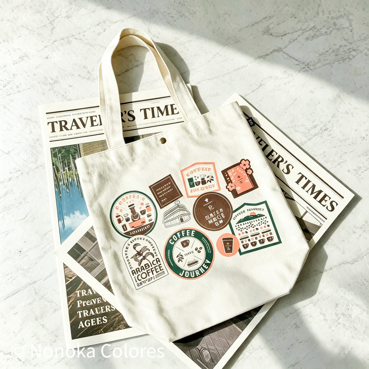 A cream canvas tote bag with multiple coffee-themed patch prints, styled on top of vintage travel magazines on a light marble surface.