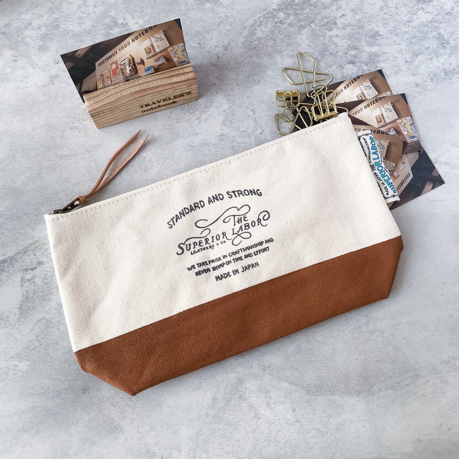Front view of The Superior Labor brown canvas pouch laid next to its original fabric packaging bag.