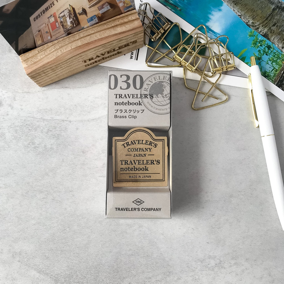 A boxed Traveler's Notebook brass clip set on a light gray stone surface, surrounded by desk props including a pen, paper clips, and a wooden block.