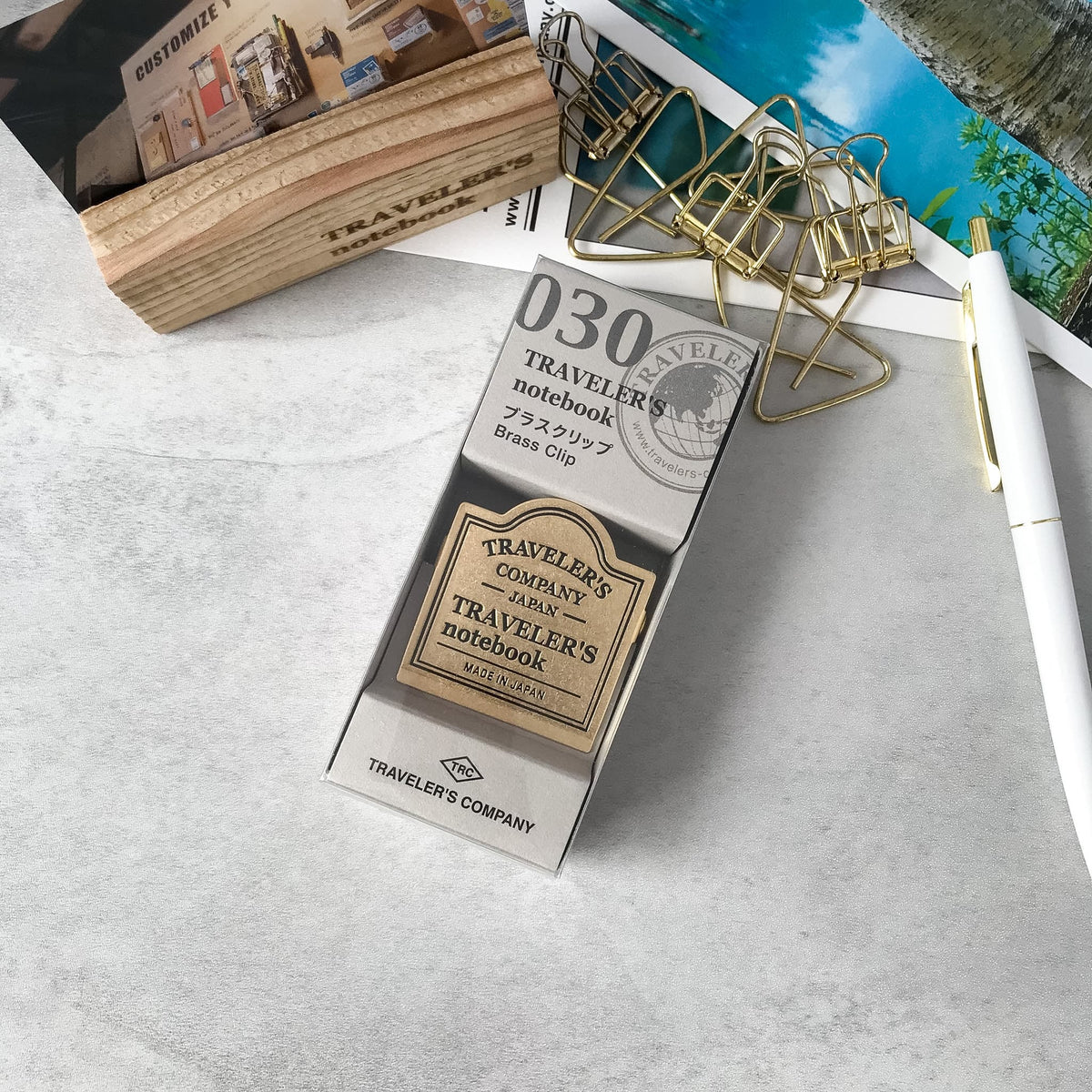 A boxed brass clip labeled 'D30 Traveler's notebook French clip' photographed on a light gray surface with styled desk props including a pen, paper clips, and a notebook.