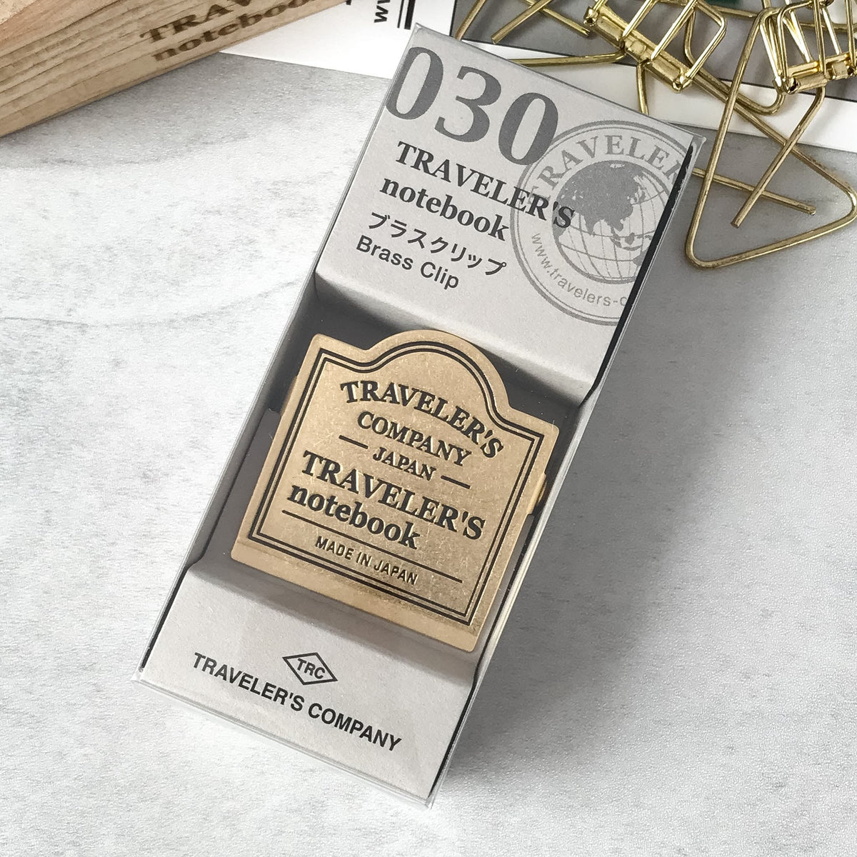 Traveler's Notebook brass clip in its original packaging, displayed on a light gray marble surface with gold paperclips in the background.