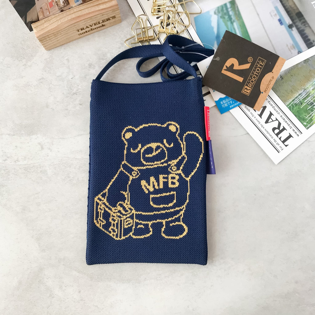 A navy blue woven crossbody bag with a cartoon bear holding a suitcase and the letters 'MFB' on the front, styled on a light gray surface with travel-themed props.
