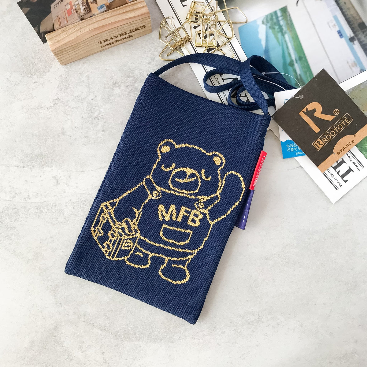 A navy blue knitted pencil pouch featuring a gold embroidered bear character holding a suitcase with 'MFB' on the chest. The pouch is styled on a light gray marble surface with office accessories, a paper tag reading 'MOONOTE', and a wooden block.