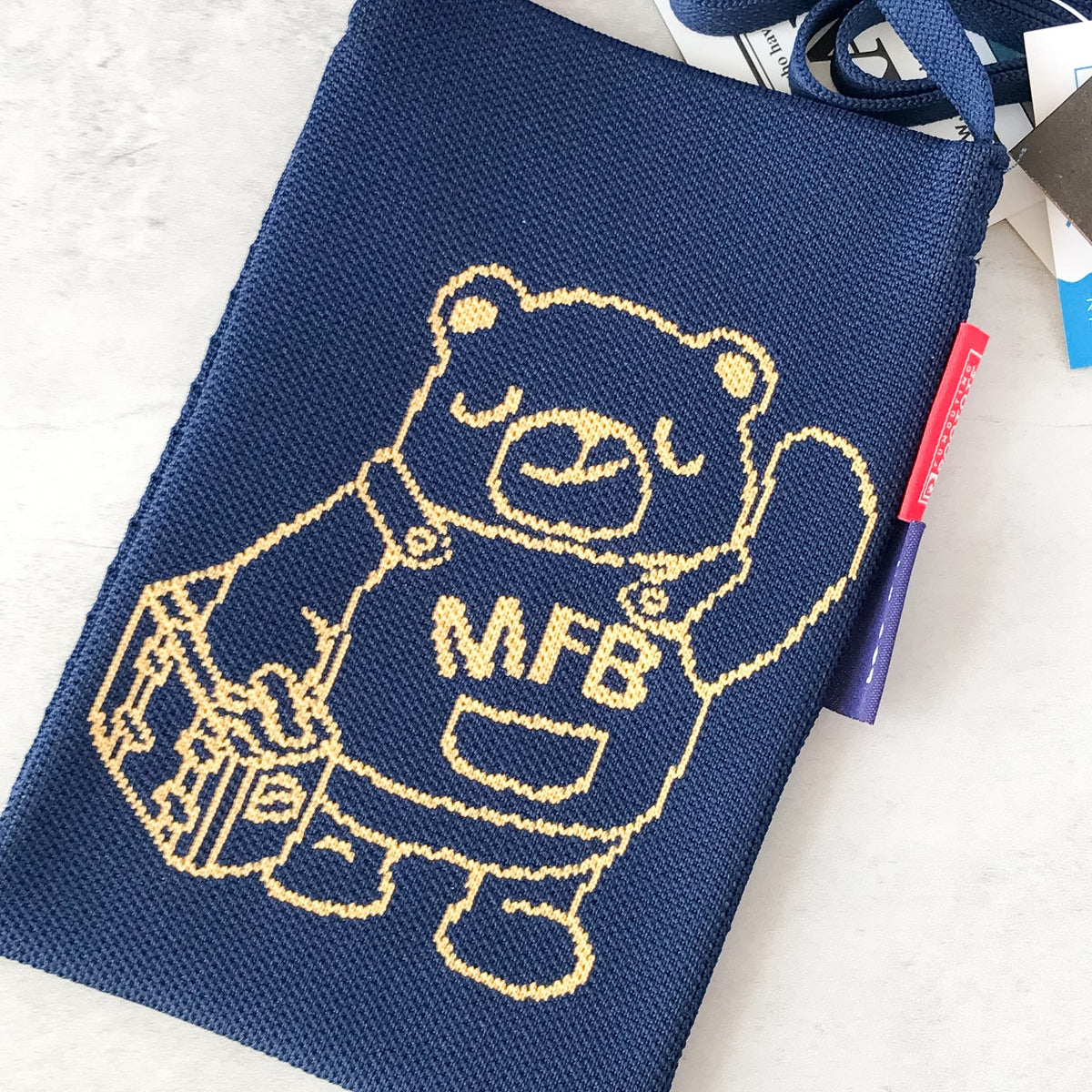 A navy blue woven fabric pouch featuring a cream-colored embroidered cartoon bear wearing overalls with 'MFB' on the chest, shown on a light textured surface.