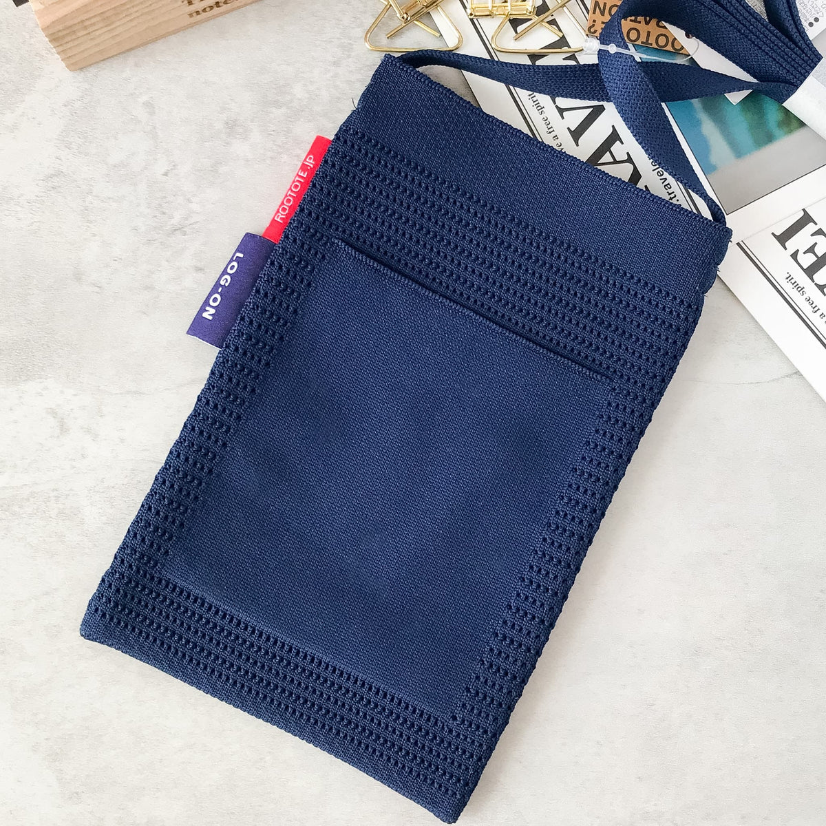 A navy blue knit fabric pouch or sleeve with a small pocket, laid flat on a light surface with a few styled paper props around it.