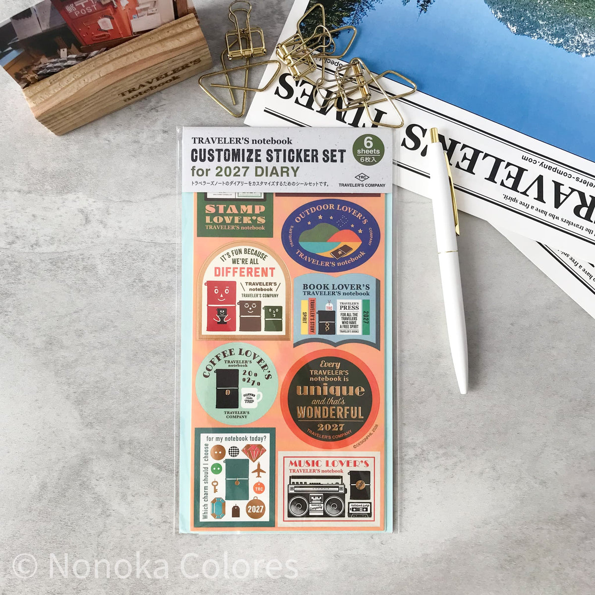 A sheet of customizable planner stickers for a 2027 diary by Nonoka Colores, styled in a flat-lay with a white pen, gold paperclips, and a wooden stamp block on a light gray marble surface.