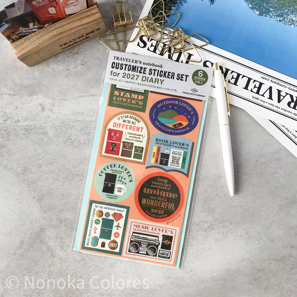 A sheet of custom sticker set for 2027 by Traveler's Notebook, styled on a gray marble desk with gold paperclips, a white pen, and a wooden block.