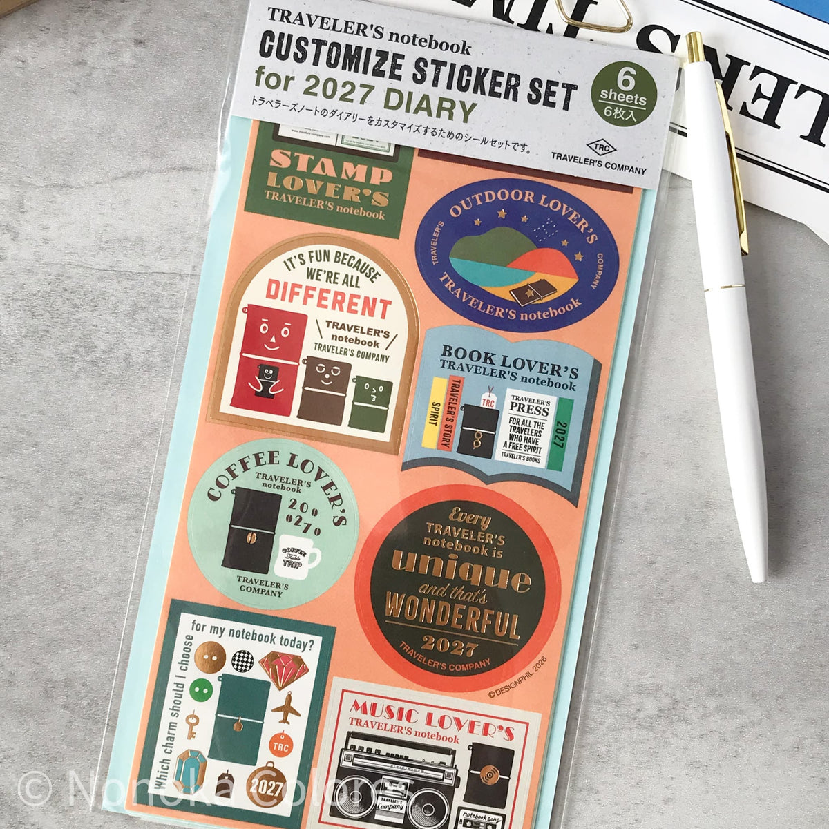 A packaged 6-piece custom sticker set for a 2027 diary, featuring various travel-themed stickers like 'Coffee Lover's', 'Outdoor Lover's', and 'Music Lover's' on a peach and light blue backing card, styled on a gray textured surface with a white pen.