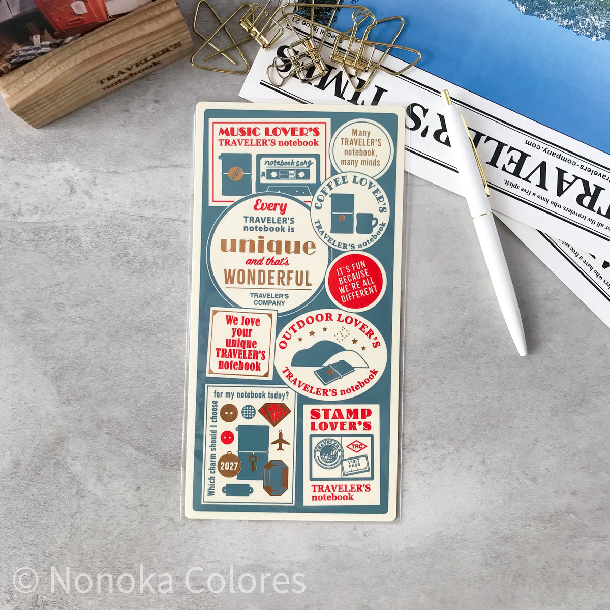 A rectangular sticker sheet with vintage-style travel-themed labels and quotes, placed on a gray surface with a white pen, binder clips, and printed papers around it. The sheet includes text such as 'Music Lover's Notebook' and 'Every traveler's notebook is unique and worth wonderful.