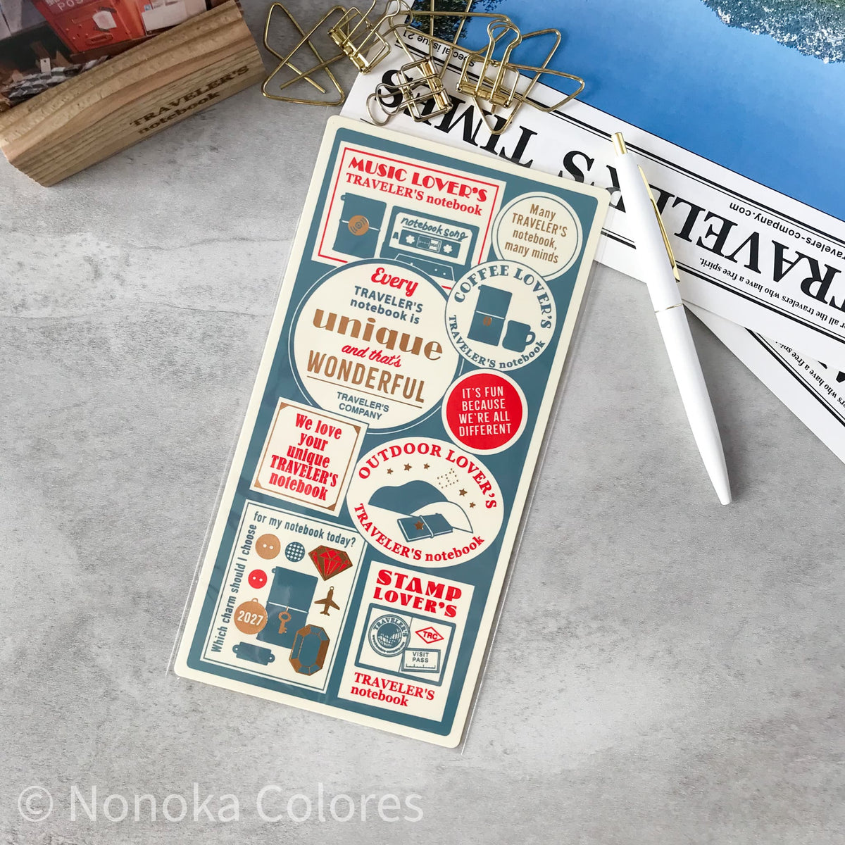 A rectangular notebook or planner cover with a vintage travel-themed sticker collage, photographed on a light gray surface with clips, a pen, and newspaper around it. The cover includes phrases like 'Music Lover's Traveler's Notebook' and 'Every traveler's hometown is unique and wonderful.