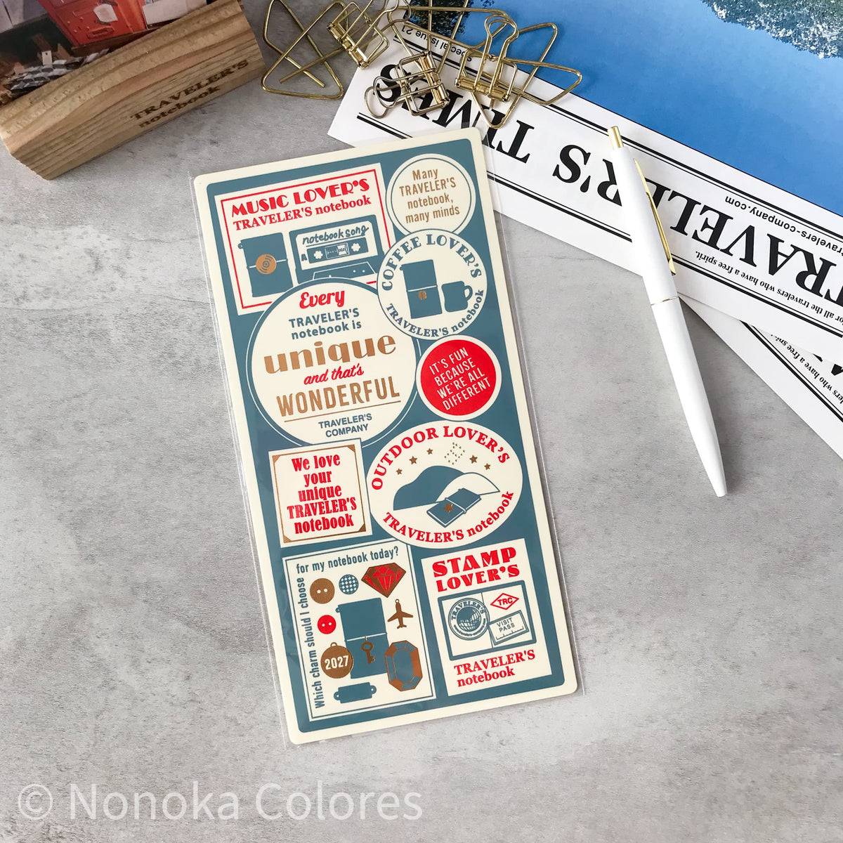 A rectangular notebook or journal cover with vintage travel-themed sticker designs, photographed on a light gray surface with a white pen, binder clips, and printed papers around it. The cover includes phrases like 'Music Lover's Traveler's Notebook' and 'Every traveler's notebook is unique and wonderful.