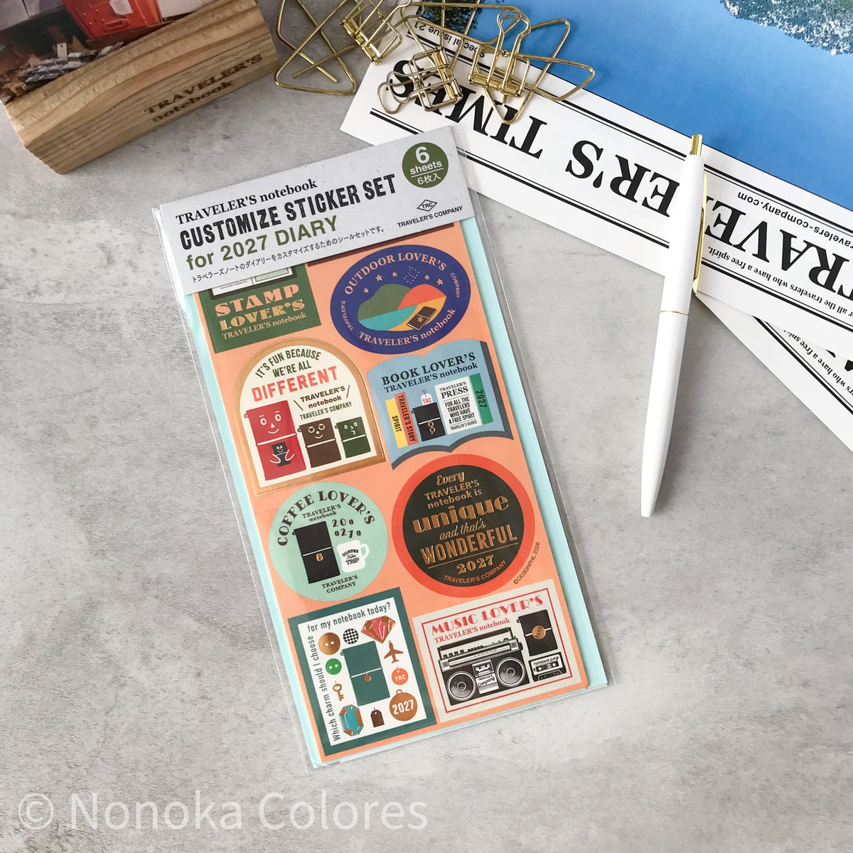 A packaged sticker set for a 2027 diary/traveler notebook, shown on a light gray surface with a gold wire clip, a white pen, and printed papers in the background. The sticker sheet contains multiple retro-style labels and stickers in pastel and bright colors, including phrases like 'Start Love,' 'Book Lover,' 'Coffee Lover,' and 'Music Lover.