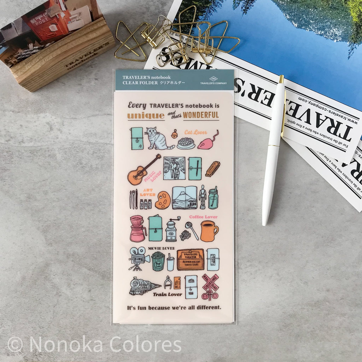 A sheet of Nonoka Colors Traveler's Notebook Clear Filler stickers displayed on a gray stone surface with stationery props including a white pen, gold wire clips, a wooden stamp block, and printed papers.