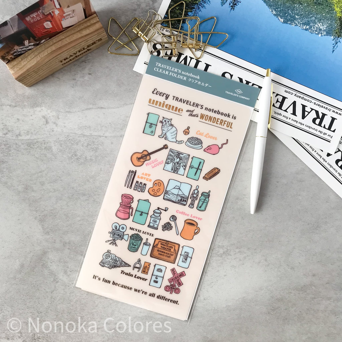 A rectangular sticker sheet featuring travel-themed illustrations and quotes, styled on a gray textured surface with a gold binder clip, white pen, and printed papers in the background. The sticker sheet has a pale pink base with multicolored drawings and text including 'Every traveler's notebook is wonderful' and 'It's fun because we're all different.