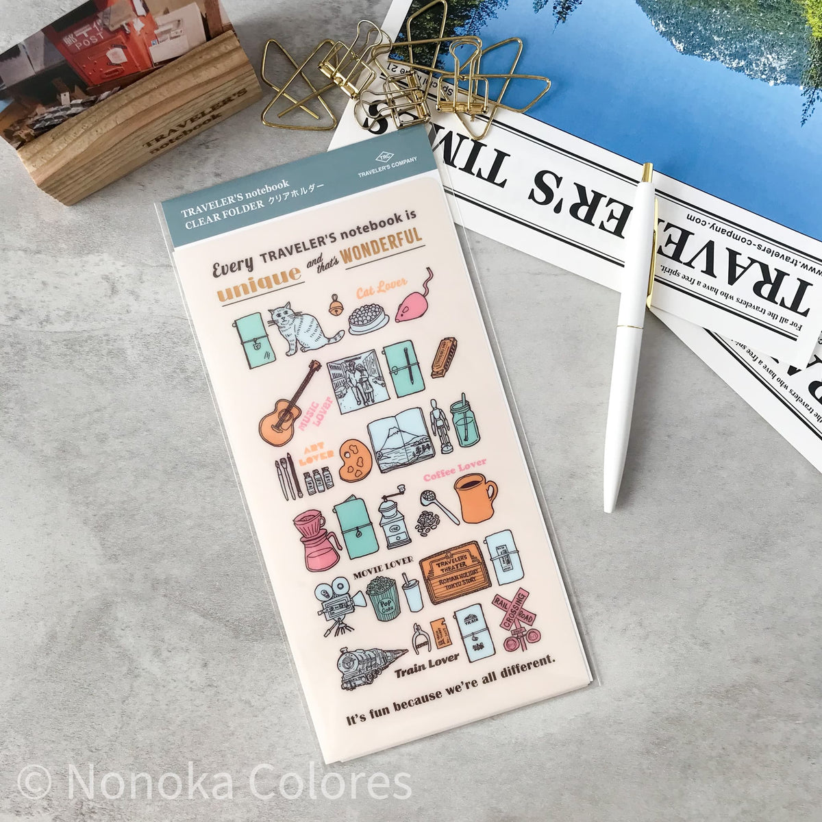 A sheet of Nonoka Colores 'Traveler's Notebook' decorative stickers displayed on a gray tabletop with a white pen, gold binder clips, and a magazine.