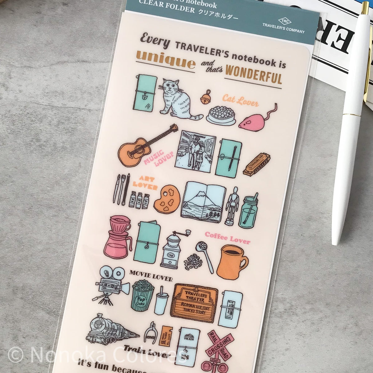 A clear folder sticker sheet from Trade Line Company featuring cute travel-themed illustrations like a cat, guitar, coffee maker, and train, with text such as 'Every TRAVELER'S notebook is unique and that's WONDERFUL'. The sheet is displayed on a gray textured surface next to a white pen.
