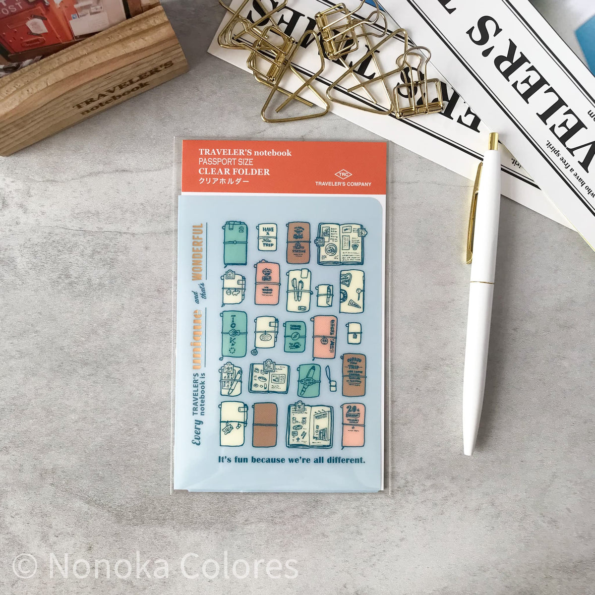 A clear planner folder sticker sheet from Traveler's Notebook, featuring various illustrated travel luggage and suitcase designs in pastel colors. The sheet is displayed on a gray textured desk alongside a white pen, gold paperclips, and a wooden stamp block.