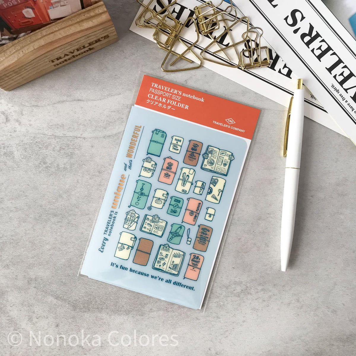 A sheet of Nonoka Colores Traveler's notebook clear folder stickers displayed on a gray textured surface next to a white pen and gold paperclips.