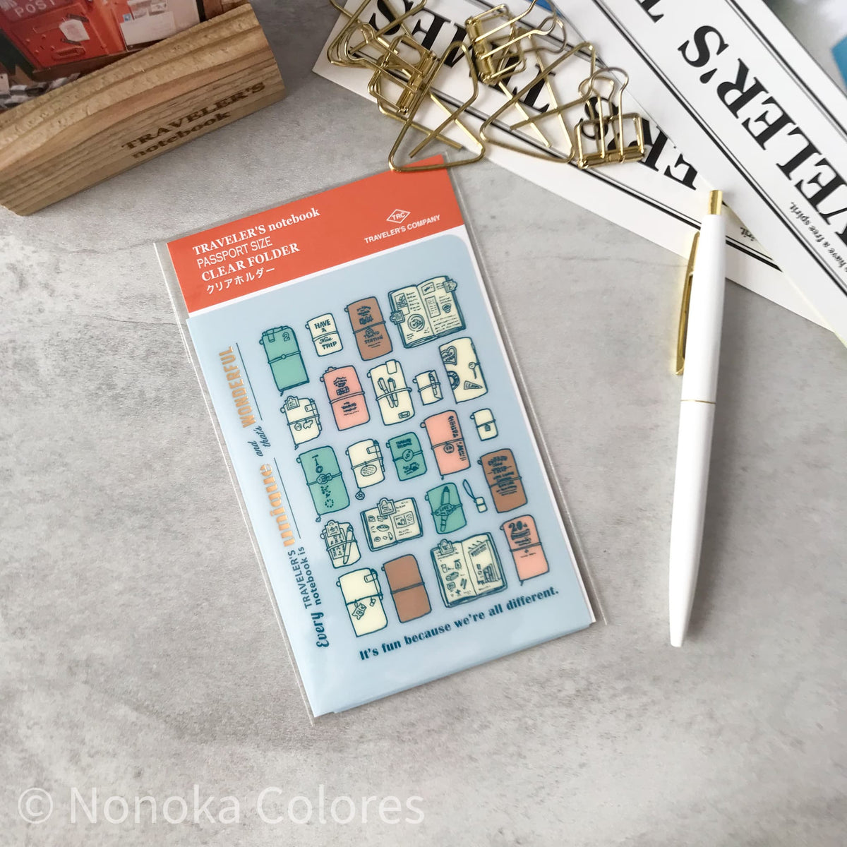 A packaged clear stamp set labeled Traveler's Notebook Clear Sticker, shown on a light gray tabletop with stationery props including a white pen, gold paper clips, and printed papers. The stamp sheet has multiple illustrated luggage and travel-themed sticker designs in muted teal, peach, tan, and cream on a pale blue background.
