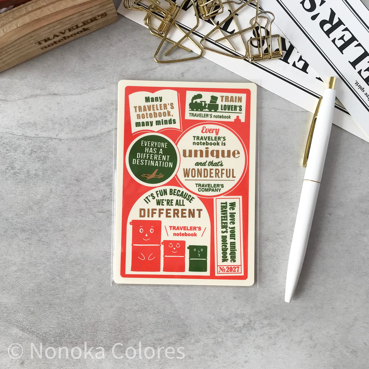 A rectangular notebook cover or notebook card with retro travel-themed stickers and text, photographed on a light gray surface with a white pen, gold binder clips, and printed papers around it. The product itself is cream with red, white, and dark green printed graphics and text.