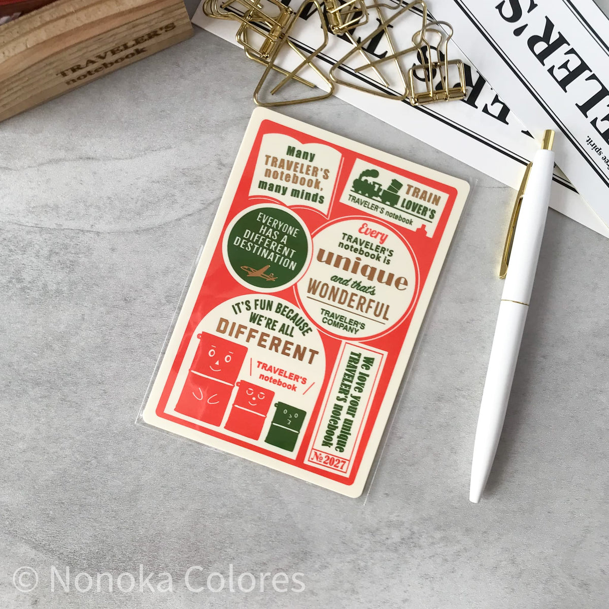 A rectangular sticker sheet with retro travel-themed illustrations and text, placed on a light gray surface with a white pen, gold paperclips, and printed papers around it.