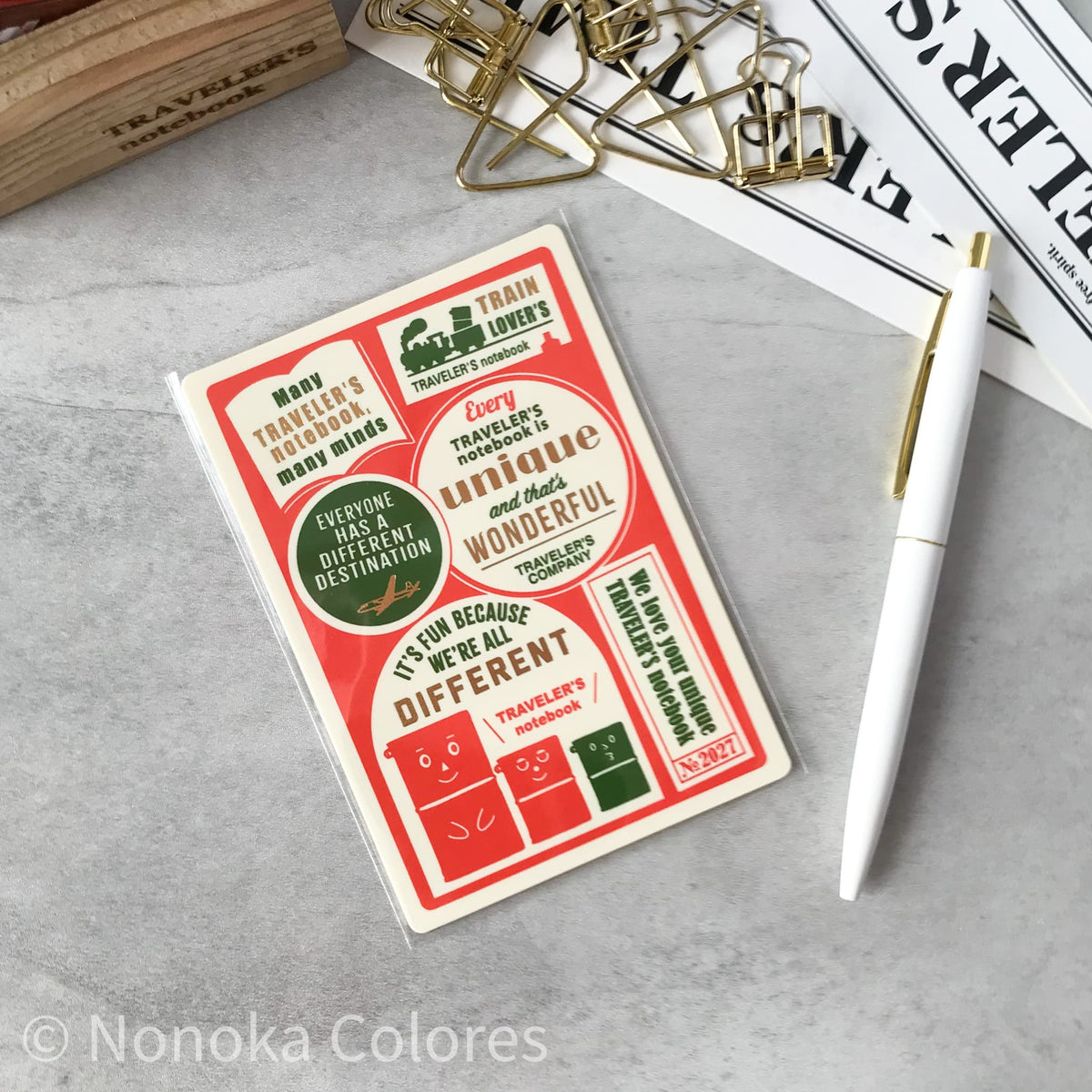 A rectangular sticker sheet with vintage-style red, green, and cream travel-themed graphics and text, photographed on a light gray surface with a white pen, gold paper clips, and printed papers nearby.