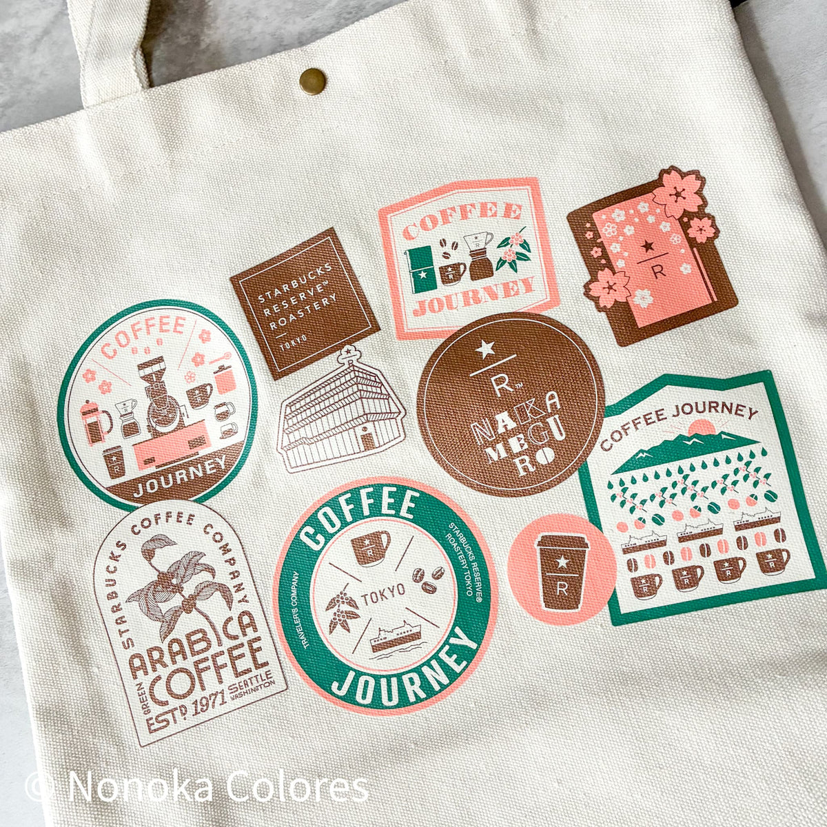 An off-white canvas tote bag featuring multiple colorful coffee-themed graphic prints in pink, green, and brown, laid flat on a light gray surface.