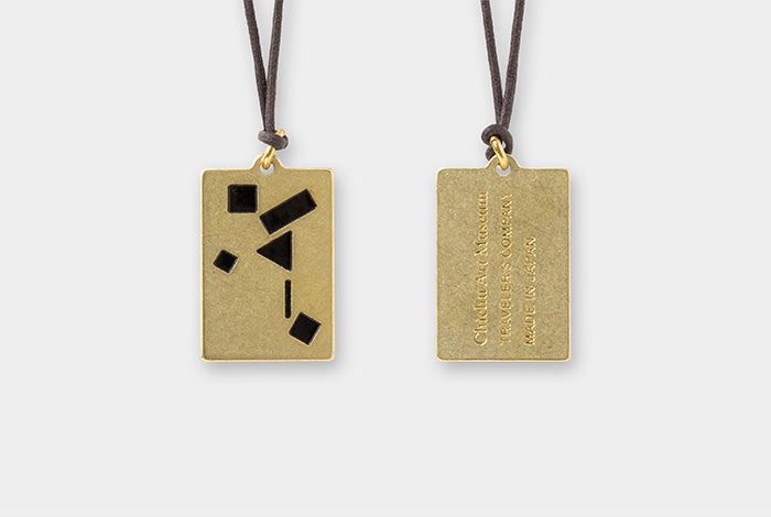 Two rectangular gold-toned pendant necklaces on dark cords, shown against a light gray background. One pendant features black geometric inlays, and the other shows engraved text.