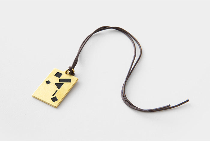 A gold rectangular pendant with black geometric inlay details, suspended from a dark brown leather cord, photographed on a light gray background.
