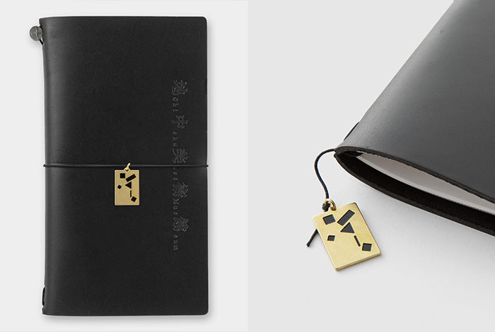A black leather journal with a wrap-around strap and a small gold geometric tag, shown in a split view: one full-length shot on the left and a close-up on the right against a light gray background.