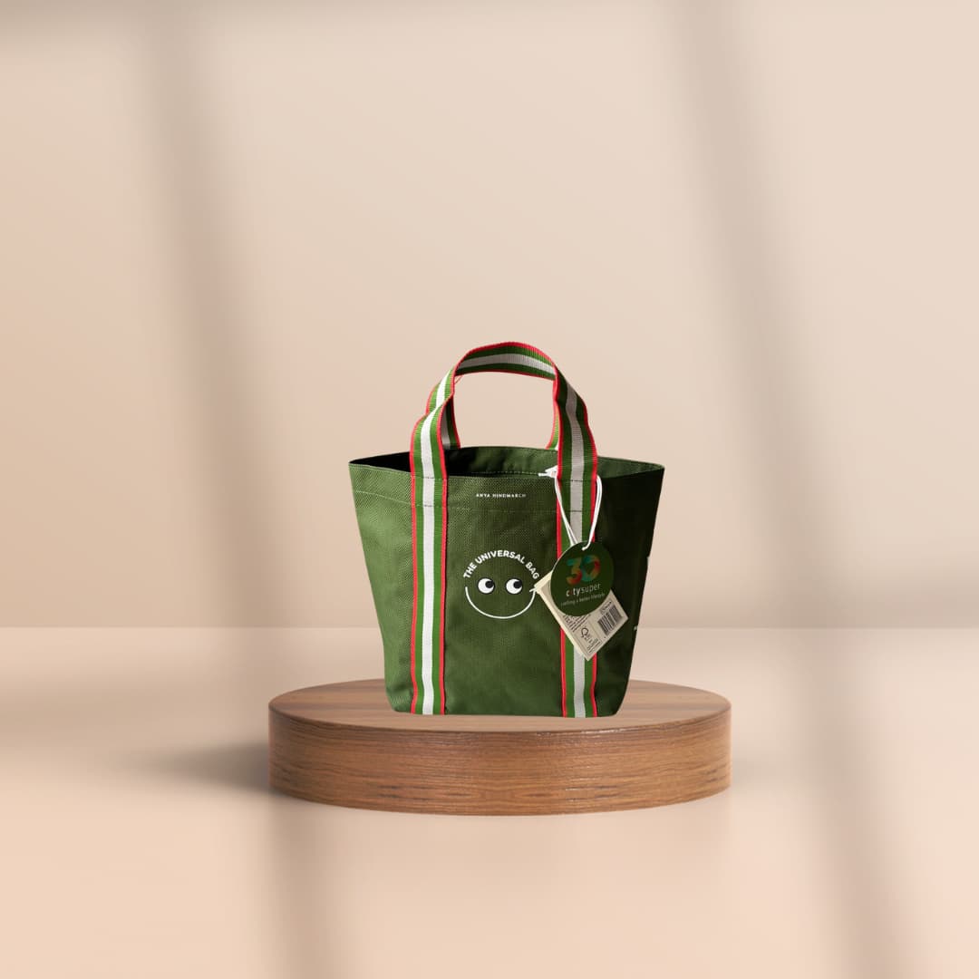 A green fabric tote bag with striped handles and a hanging tag, displayed on a round wooden pedestal against a warm beige background.