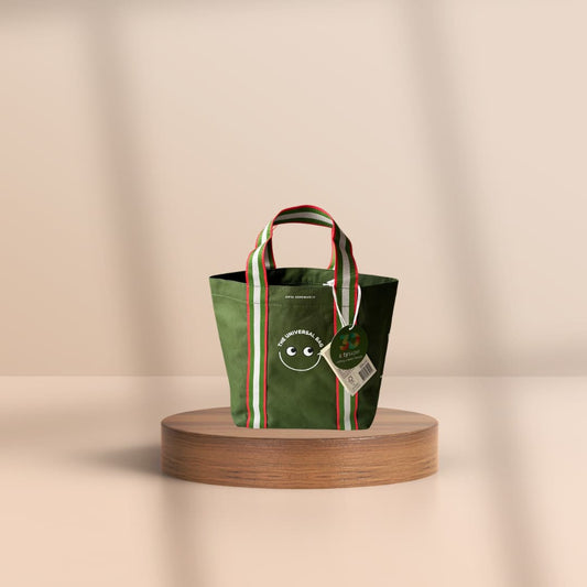 A green fabric tote bag with striped handles and a hanging tag, displayed on a round wooden pedestal against a warm beige background.