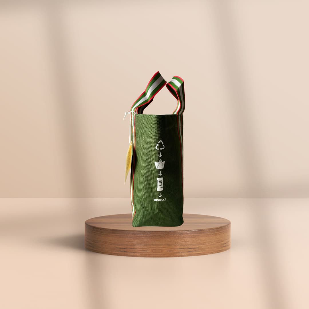 A green reusable tote bag with dual handles, featuring a white recycling graphic and the word 'RECYCLE' on the front, displayed on a round wooden pedestal against a warm beige background.