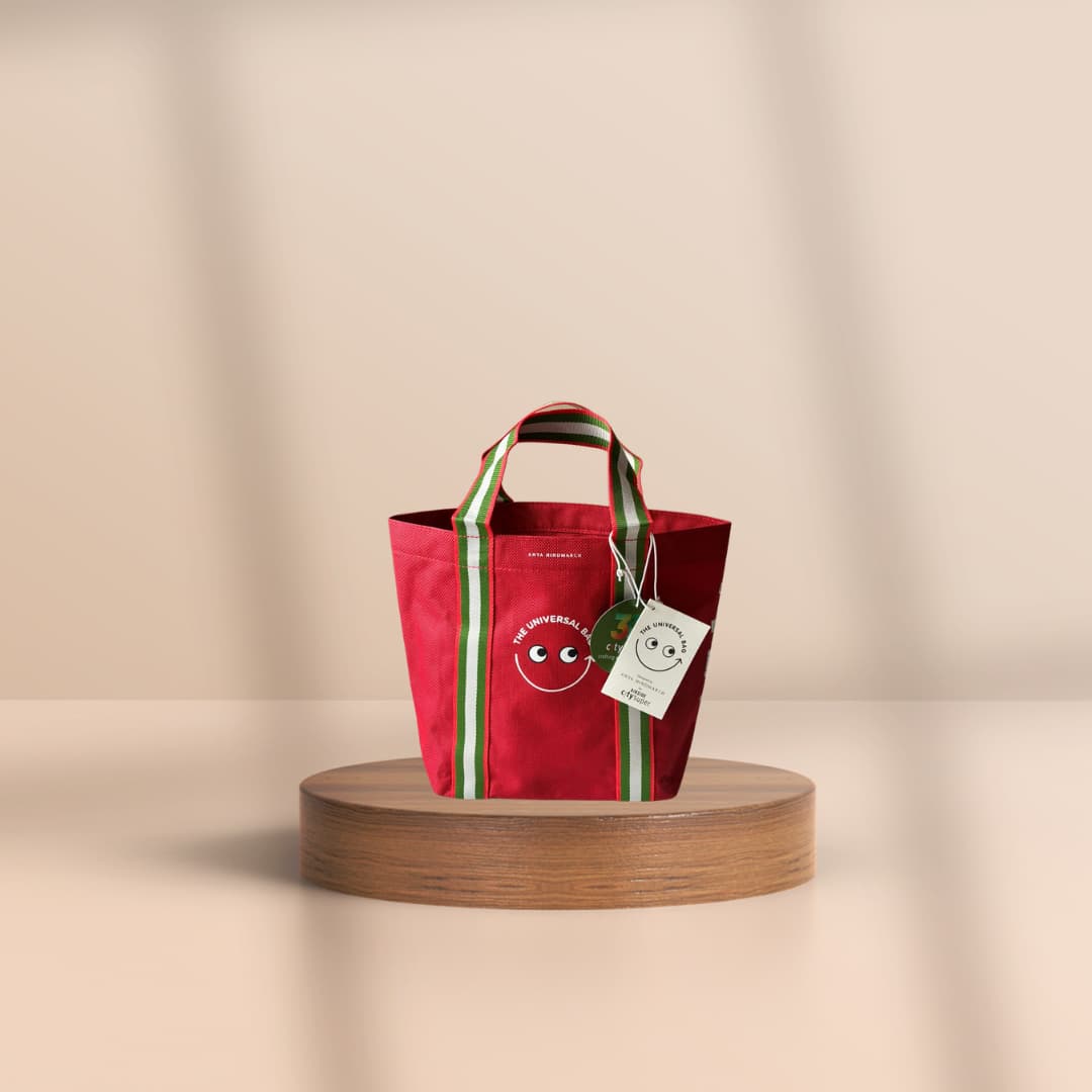 A red fabric tote bag with green and white striped handles and side stripes, featuring a cartoon face graphic and a hanging tag, displayed on a round wooden pedestal against a warm beige background.