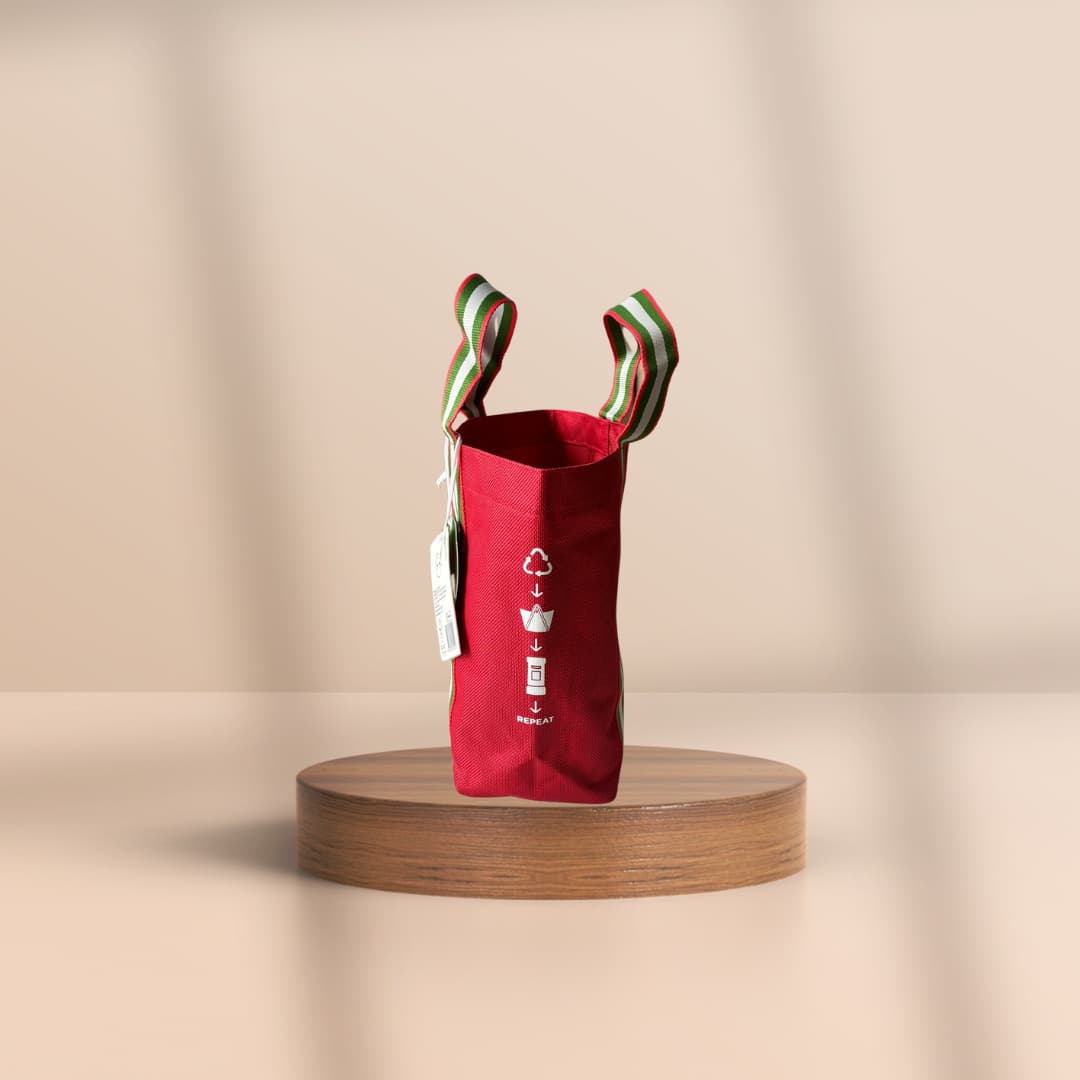 A red fabric tote bag with striped handles, displayed on a round wooden pedestal against a warm beige background.