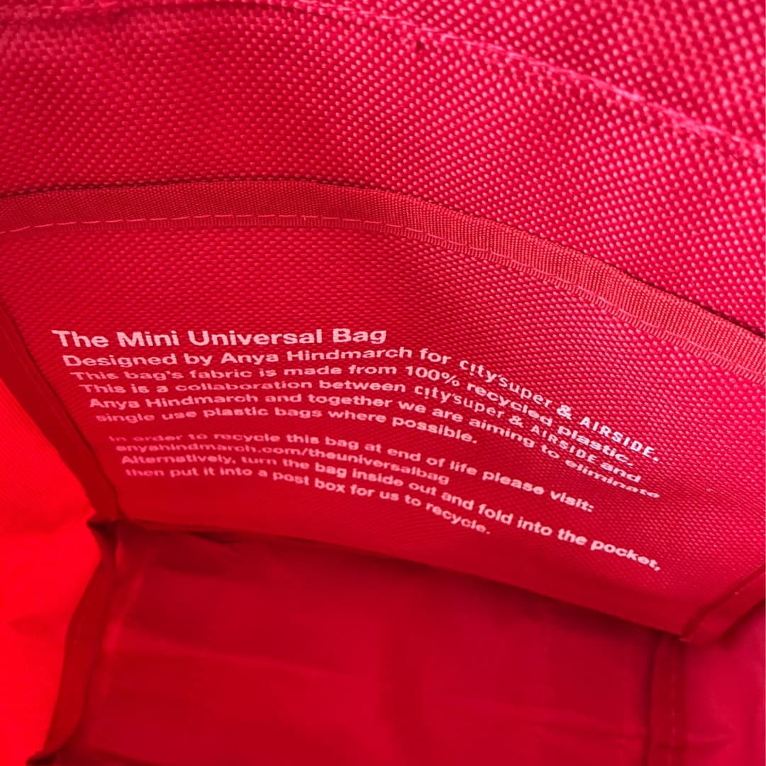 Close-up of a red fabric interior showing printed white text that reads 'The Mini Universal Bag' and recycling information for a bag made from recycled plastic.