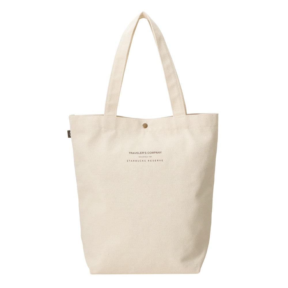 An off-white canvas tote bag with dual shoulder straps, featuring a small metal snap button closure at the top and 'TRAVELER'S COMPANY' and 'STARBUCKS RESERVE' printed in dark brown text on the front.
