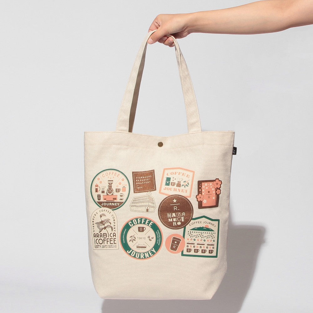An off-white canvas tote bag with a hand holding its top handles, decorated with various retro-style coffee-themed stickers in muted green, brown, and orange tones. The background is a clean, neutral light gray studio backdrop.